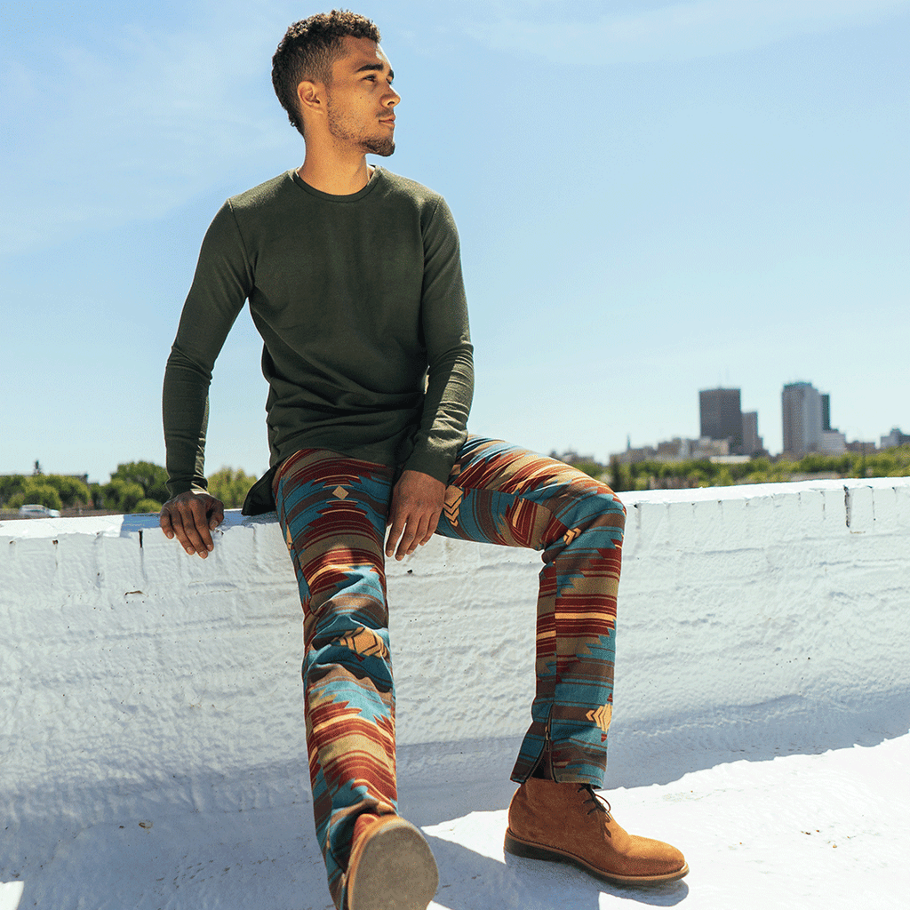 Man in green long sleeve and blue, brown, red and yellow patterned pants sitting on white brick roof overlooking Winnipeg downtown skyline.