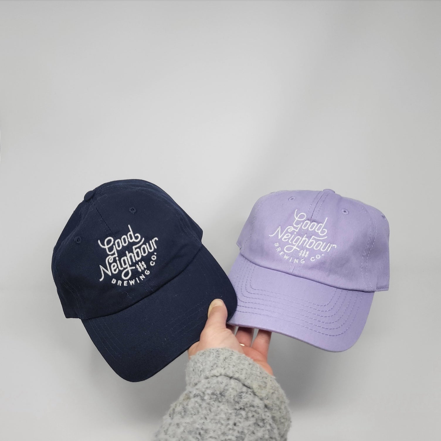 Hand holding two Navy and Light Purple Good Neighbour Brewing Co. caps on white background