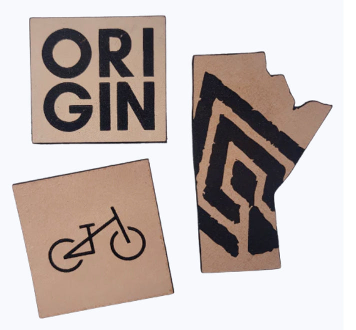 Tan and black Origin, bike graphic and Manitoba shaped Lazer cut leather patches on a white background
