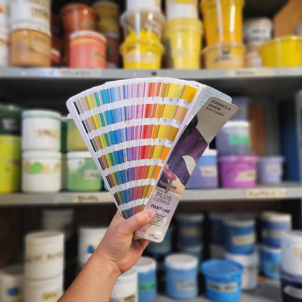 Handing holding up Pantone colour swatch booklet with screen printing ink on shelves in the background.