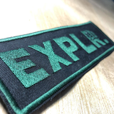 Green EXPLR. embroidered tag on black fabric.