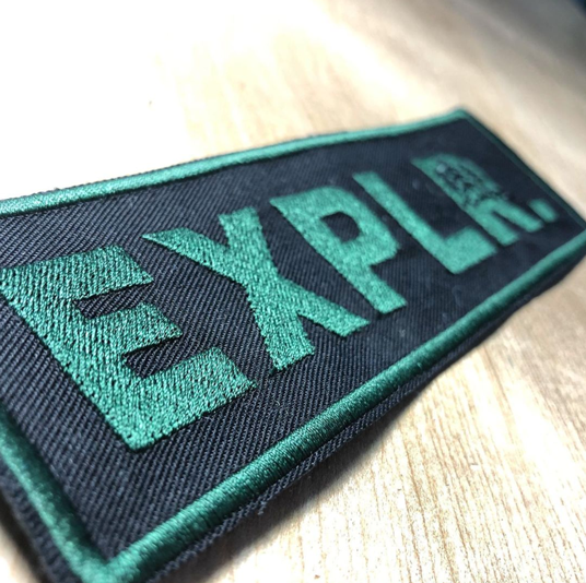 Green EXPLR. embroidered tag on black fabric.